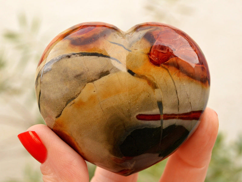 Large polychrome jasper heart crystal 70mm on hand with background with  green plants