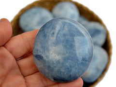 One blue calcite palm stone 70mm on hand with background with some stones inside a basket