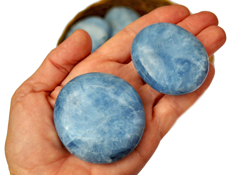 Two blue calcite palm stones 40mm-70mm on hand with background with some crystals inside a basket on white