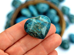 One blue apatite heart stone 30mm on hand with background with some stones inside and outside a bowl