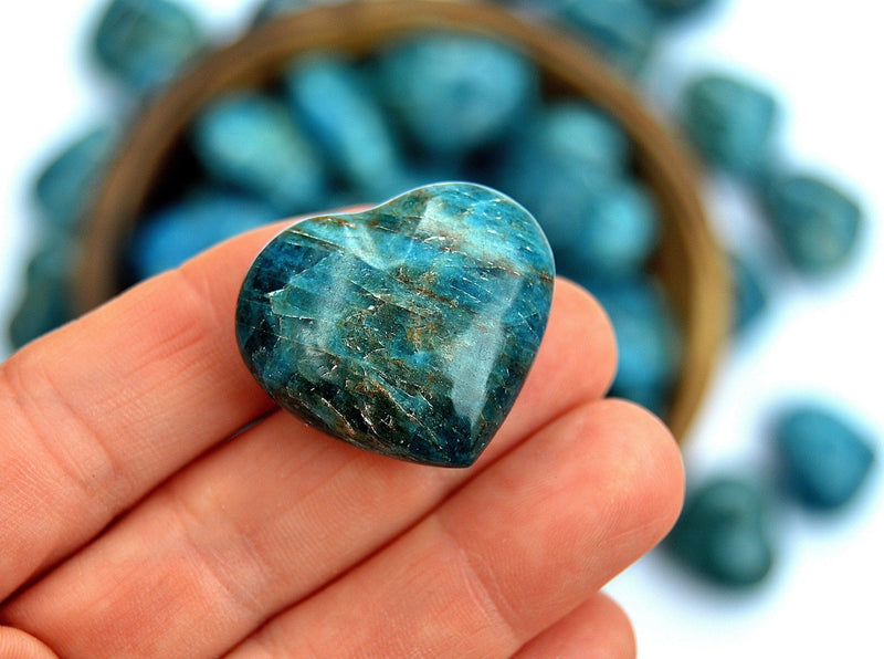 One small blue apatite heart crystal on hand with background with several hearts inside a bowl and outside