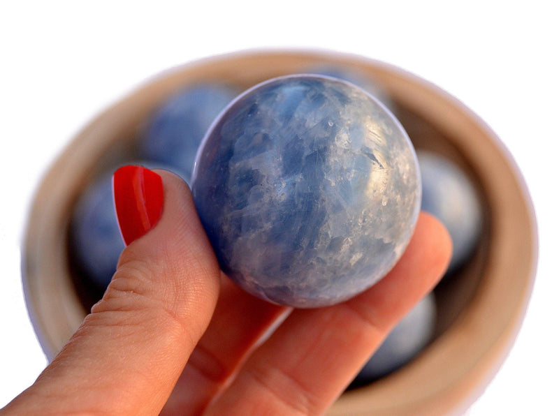 One blue calcite crystal ball 40mm on hand with background with some crystals inside a wood bowl