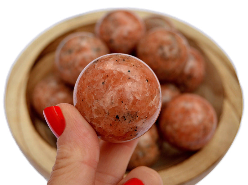 One orange calcite sphere crystal 30mm on hand with background with some balls inside a wood bowl on white