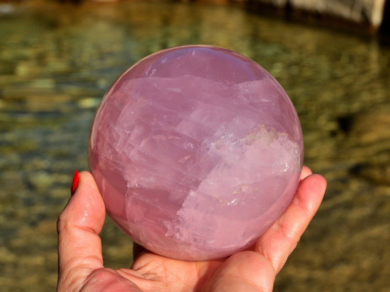 One large rose quartz sphere crystal 85mm on hand with background river landscape