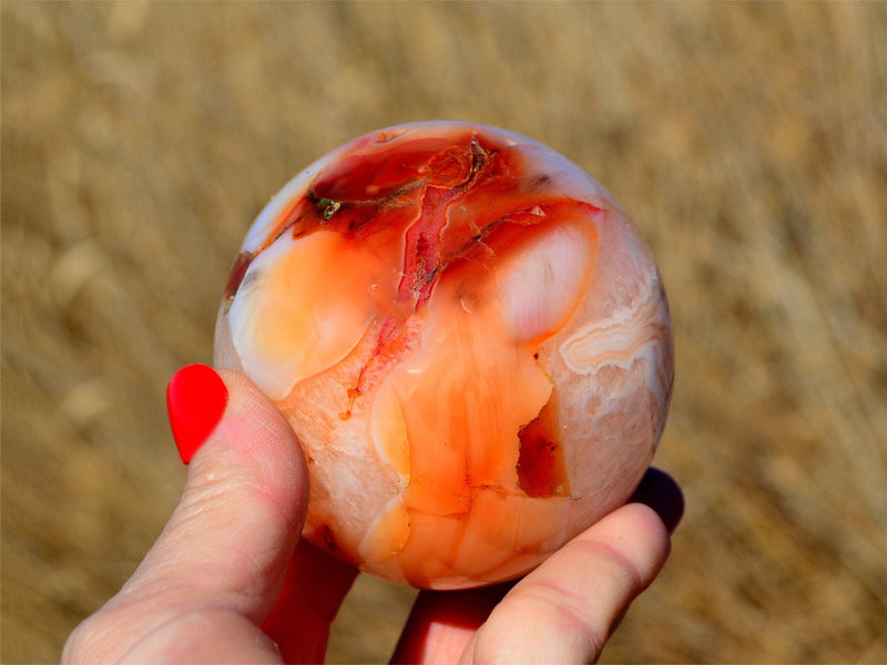 One natural carnelian sphere crystal 70mm on hand with straw landscape background