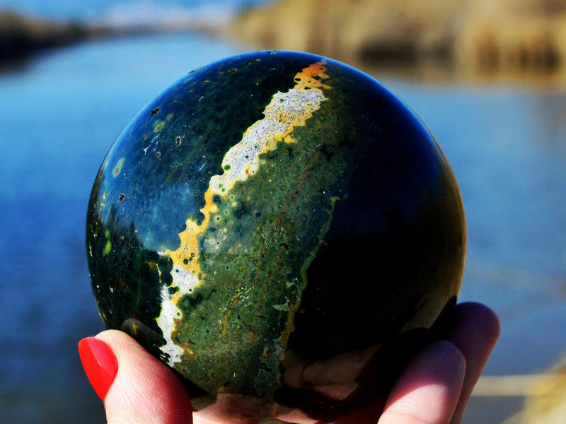 One green ocean jasper mineral sphere 90mm on hand with river landscape background