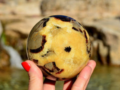 Large yellow septarian sphere crystal 90mm on hand with river landscape background