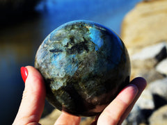 One blue labradorite crystal sphere 75mm on hand with background with river and rocks landscape